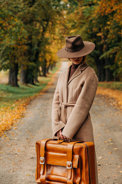 Европейская женщина в коричневом пальто и шляпе 1940-х годов ходит по тропинке через деревья с ярко-желтой осенней листвой