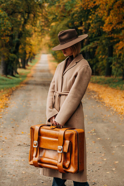 Европейская женщина в коричневом пальто и шляпе 1940-х годов ходит по тропинке через деревья с ярко-желтой осенней листвой