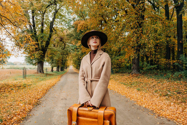 Европейская женщина в коричневом пальто и шляпе 1940-х годов ходит по тропинке через деревья с ярко-желтой осенней листвой