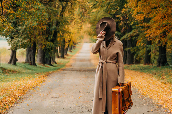 Европейская женщина в коричневом пальто и шляпе 1940-х годов ходит по тропинке через деревья с ярко-желтой осенней листвой