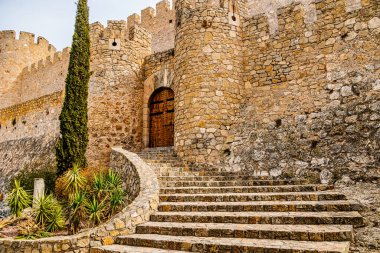 Görüntü, eğri bir merdivenin tepesinde büyük ahşap bir kapısı olan heybetli bir taş kale yakalıyor.