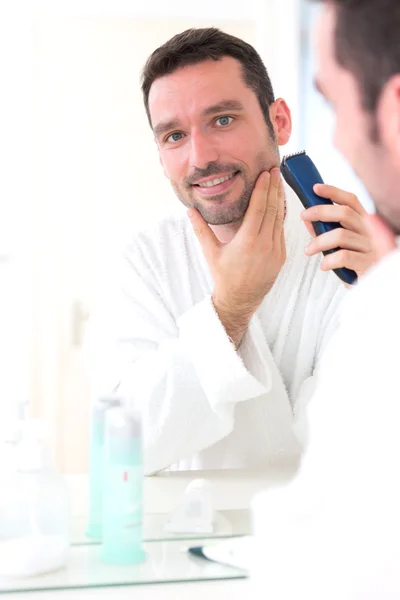 Jovem homem atraente barbeando sua barba na frente de um espelho — Fotografia de Stock