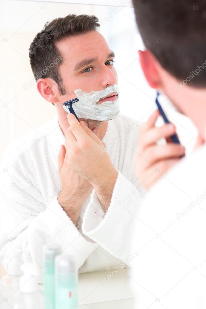 Young attractive man shaving his beard in front of a mirror