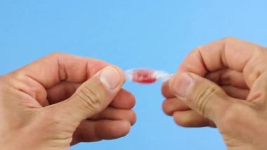 Eller, şeffaf bir ambalaj kağıdından, mavi arka plandan, sağlıksız şeker şekerlerinden ve plastik çöpten çıkan kırmızı şeker lolipopunu açar.