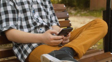 Akıllı telefon tutan bir çocuğun ellerini kapat. Çocuk dışarıda bankta oturuyor. Şehir parkı, yaz günü.