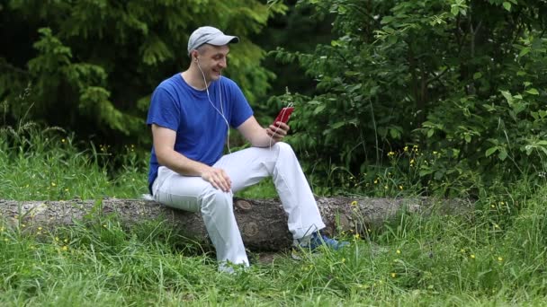 Człowiek z czerwonym smartphone siedzi na spasionego drzewa w lesie — Wideo stockowe