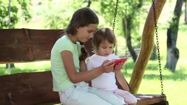 Дві красиві дівчата з червоним смартфоном сидять на гойдалці в саду — стокове відео