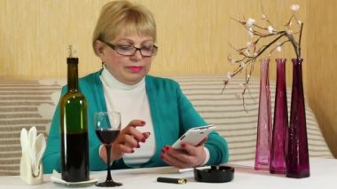 Kadın masada oturur, kırmızı şarap içiyor ve smartphone üzerinde görüşmeler