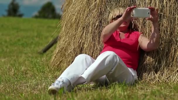 Mujer rubia se sienta en la hierba verde y hace fotos en el teléfono inteligente — Vídeos de Stock