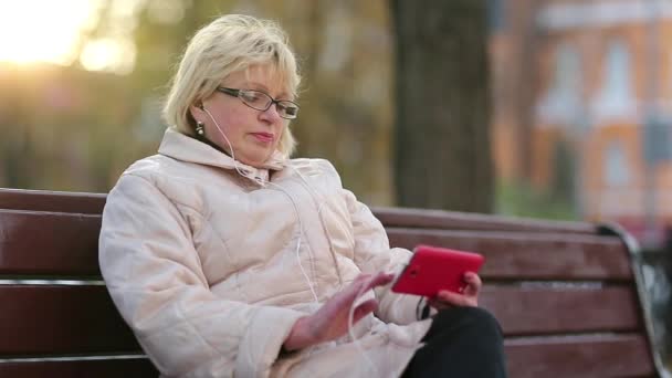 Woman sits on the bench near the road and uses red cell phone — Stock Video