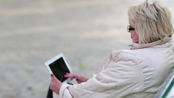 Женщина сидит на скамейке и пользуется планшетным компьютером — стоковое видео