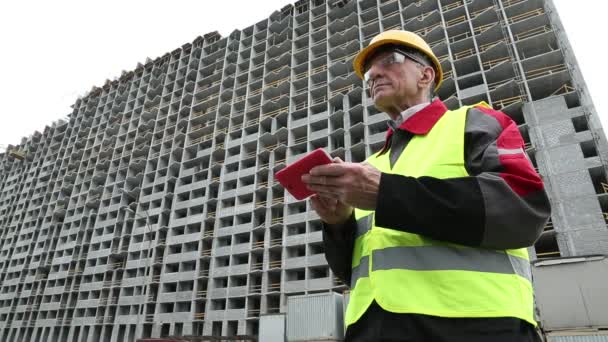 Инженер в желтом шлеме стоит с красным смартфоном рядом с недостроенным домом — стоковое видео
