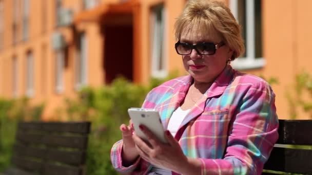 Frau sitzt auf Bank im Hof und nutzt weißes Smartphone — Stockvideo