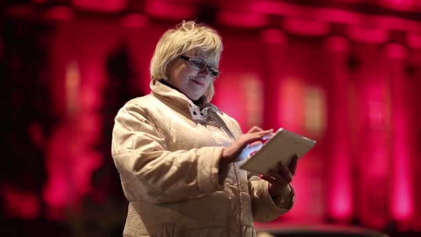 Senior vrouw staat in de buurt van rode gebouw in de nacht en maakt gebruik van Tablet Pc — Stockvideo