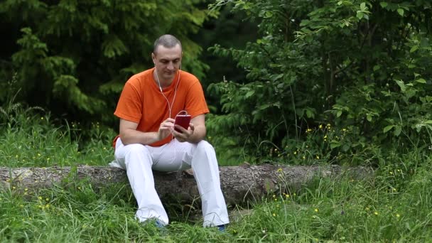 Hombre con teléfono inteligente rojo se sienta en un árbol caído en el bosque — Vídeo de stock