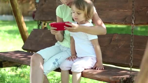 Dos chicas agradables con teléfono inteligente rojo se sienta en el banco de oscilación en el jardín — Vídeos de Stock