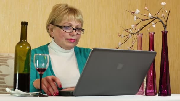 Woman with a bottle of wine sits at a table and communicates via laptop — Stock Video