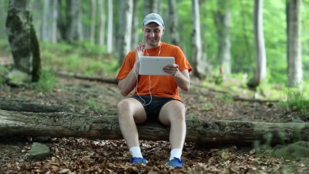 Человек сидит на упавшем дереве в лесу и общается через планшетный компьютер — стоковое видео