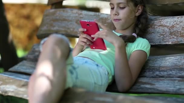 Menina bonita com smartphone vermelho senta-se no banco de balanço — Vídeo de Stock