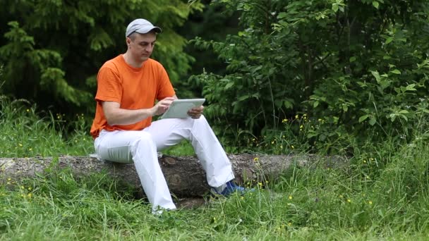 Homem com um computador tablet senta-se em uma árvore caída na floresta — Vídeo de Stock