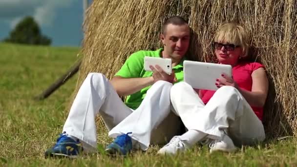 Hombre con smartphone blanco y mujer con tablet PC blanca se sienta cerca de un pajar — Vídeos de Stock