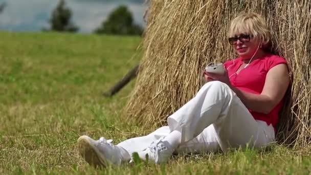 Woman sits near haystack and listens to music — Stock Video