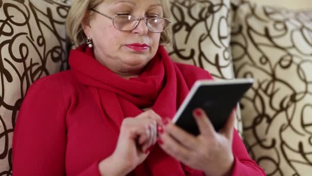 Senior woman sits on a sofa and uses electronic book. — Stock Video