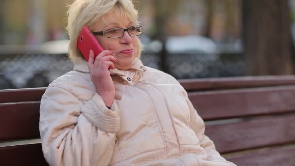 Женщина сидит на скамейке и говорит на красном смартфоне — стоковое видео