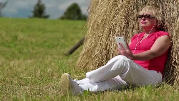 Mujer con teléfono inteligente blanco se sienta cerca de pajar y escuchar música — Vídeos de Stock