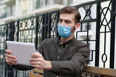Tıbbi koruyucu maskeli genç adam bankta oturup tablet bilgisayarında fotoğraf çekiyor.