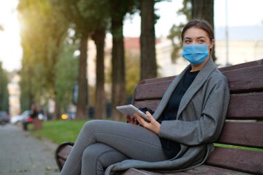 Tıbbi koruyucu yüz maskeli genç kadın sonbahar şehir meydanında bankta oturuyor ve el bilgisayarını tutuyor.