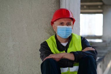 Tıbbi koruyucu yüz maskesi takan işçiler inşaat alanında oturur ve kameraya bakar.