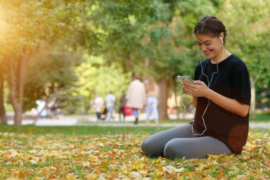 Genç güzel kız sonbahar parkında sarı yaprakların üzerinde oturur, akıllı telefon kullanır, müzik dinler ve gülümser.