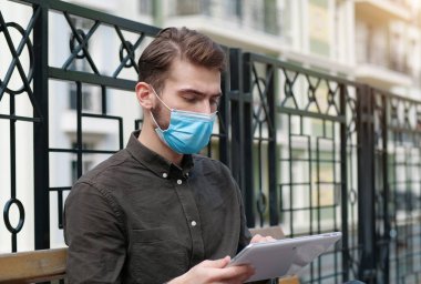 Tıbbi koruyucu yüz maskesi takan genç yakışıklı iş adamı tablet bilgisayarla çalışıyor.