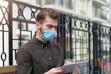 Tıbbi koruyucu yüz maskeli genç yakışıklı işadamı tablet bilgisayarla çalışıyor.