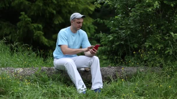 Человек сидит на бревне, держит телефон — стоковое видео