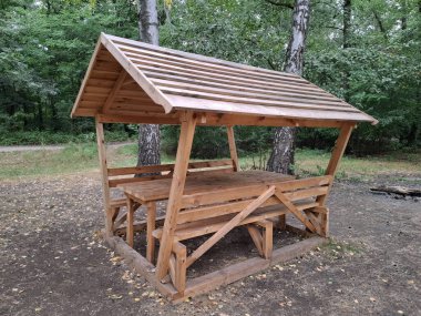 Bir orman parkında dinlenmek ve yemek yemek için yeni, ahşap bir çardak ve çatılı bir masa. Küçük bir açık hava yapısıdır ve genellikle bahçelerde veya parklarda dinlenmek için kullanılır, çatısı ve açık duvarları vardır.