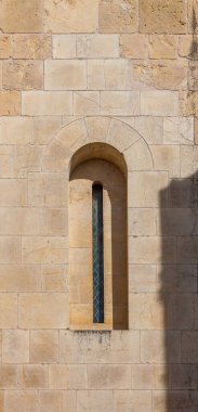 A narrow arched window with stained glass is set into a weathered stone wall, casting shadows and highlighting the texture of the ancient masonry
