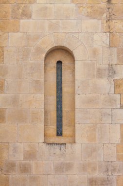 A narrow arched window with stained glass is set into a weathered stone wall, casting shadows and highlighting the texture of the ancient masonry