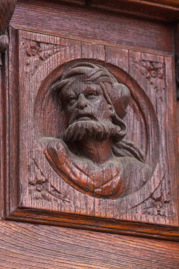 A close-up of a richly detailed, dark wood carving shows a bearded man's face with a stern expression, framed in a circular medallion on an antique wooden panel
