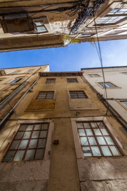 A low-angle view captures the weathered facades of tall, narrow buildings with peeling paint and grid windows, set against a clear blue sky
