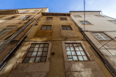 A low-angle view captures the weathered facades of tall, narrow buildings with peeling paint and grid windows, set against a clear blue sky