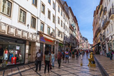 Dükkanlar ve renkli binalarla dolu hareketli bir yaya caddesinde, parlak mavi gökyüzüne karşı desenli kaldırım taşları üzerine kurulmuş, yaşayan altın bir heykeltraş yer alıyor.