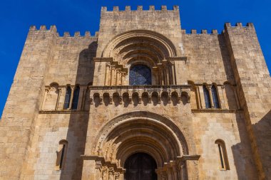 Düşük açılı bir çekim, tarihi bir taş katedralin heybetli, güneşli cephesini yakalar. Karmaşık kemerler, bir vitray pencere ve berrak mavi gökyüzüne karşı crenelleştirilmiş kuleler..