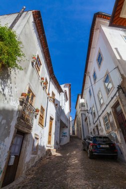 Dar bir kaldırım taşı sokak rüzgarları arasında canlı mavi gökyüzü altında uzun, beyaz yıkanmış binalar arasında modern bir araba park etmiş ve saksı bitkileri tarih sahnesine hayat katıyor.
