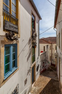 Güneşli, dar bir sokak arasında beyazlatılmış duvarlar renkli pencerelerle süslenmiş, tuhaf sokak sanatı ve parlak bir gökyüzünün altında sallanan ayakkabılar gibi seçici dekorasyonlar var..