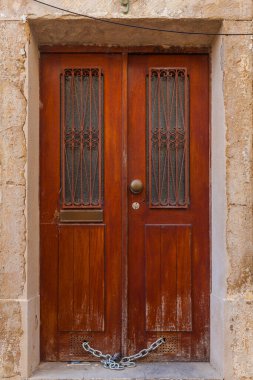 A weathered, rich brown wooden double door with ornate iron grilles is secured by a heavy chain at its base, set within a rustic stone frame