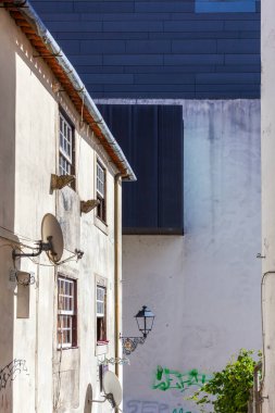 Dar bir ara sokak, parlak güneş ışığı altında çarpıcı bir kontrastlık yaratan, katı, modern koyu mavi cepheli, yıpranmış, geleneksel bir binayı yan yana getirir.