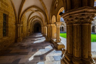Güneş ışığı kemerli açıklıklardan geçerek taştan bir zemin üzerinde büyük sütunları ve tarihi bir manastır geçidi tavanlarını aydınlatıyor.