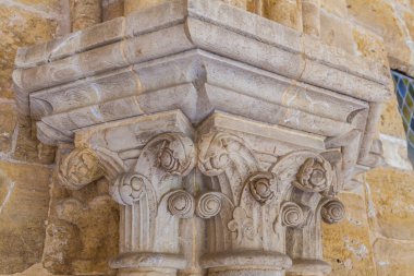 Tarihi bir binadaki sütunları destekleyen karmaşık çiçek ve kaydırma oymalarıyla süslü, yıpranmış taş bir başkentin yakın görüntüsü.